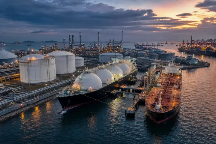 An LNG carrier and oil tanker near a busy import terminal, with storage tanks and refinery infrastructure in the background.